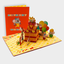 Aufgeklappte 3D Pop-Up Karte mit Geburtstagstorte, Mäusen, Ballons und Käse, daneben die zugeklappte Karte mit singender Maus und „Happy Birthday“-Schriftzug auf orangem Cover.