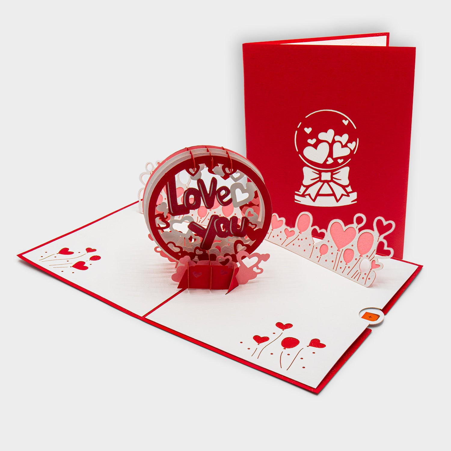 Aufgeklappte 3D Pop-Up Karte mit „Love You“-Schriftzug und Herz-Motiven, daneben die zugeklappte Karte mit Schneekugel-Design auf rotem Cover.