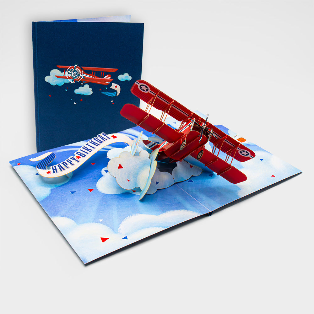 Aufgeklappte 3D Pop-Up Karte mit rotem Flugzeug und Wolken, daneben die zugeklappte Karte mit Flugzeug-Motiv auf blauem Cover.