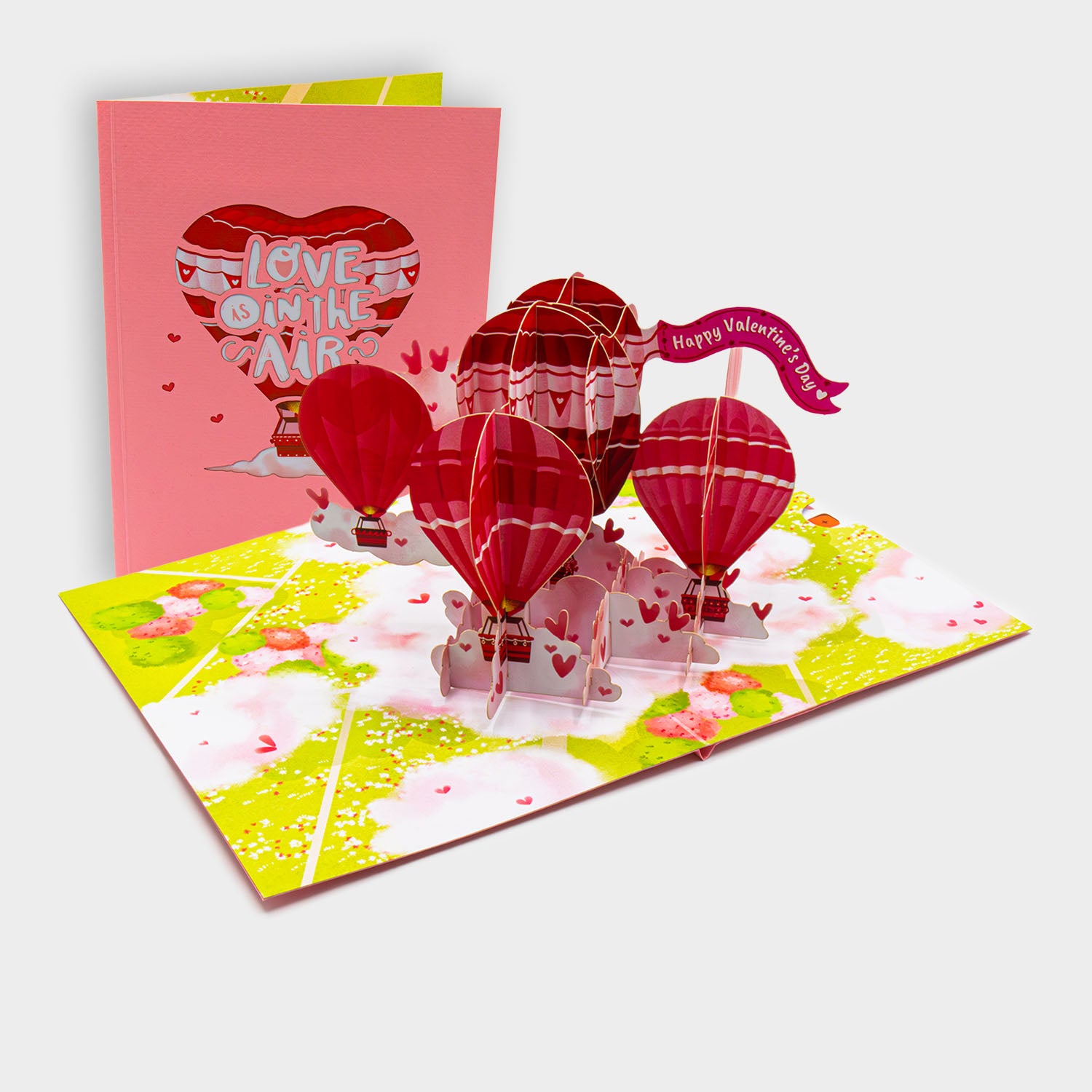 Aufgeklappte 3D Pop-Up Karte mit Heißluftballons in Herzform und Wolken, daneben die zugeklappte Karte mit „Love is in the Air“-Motiv auf rosa Cover.